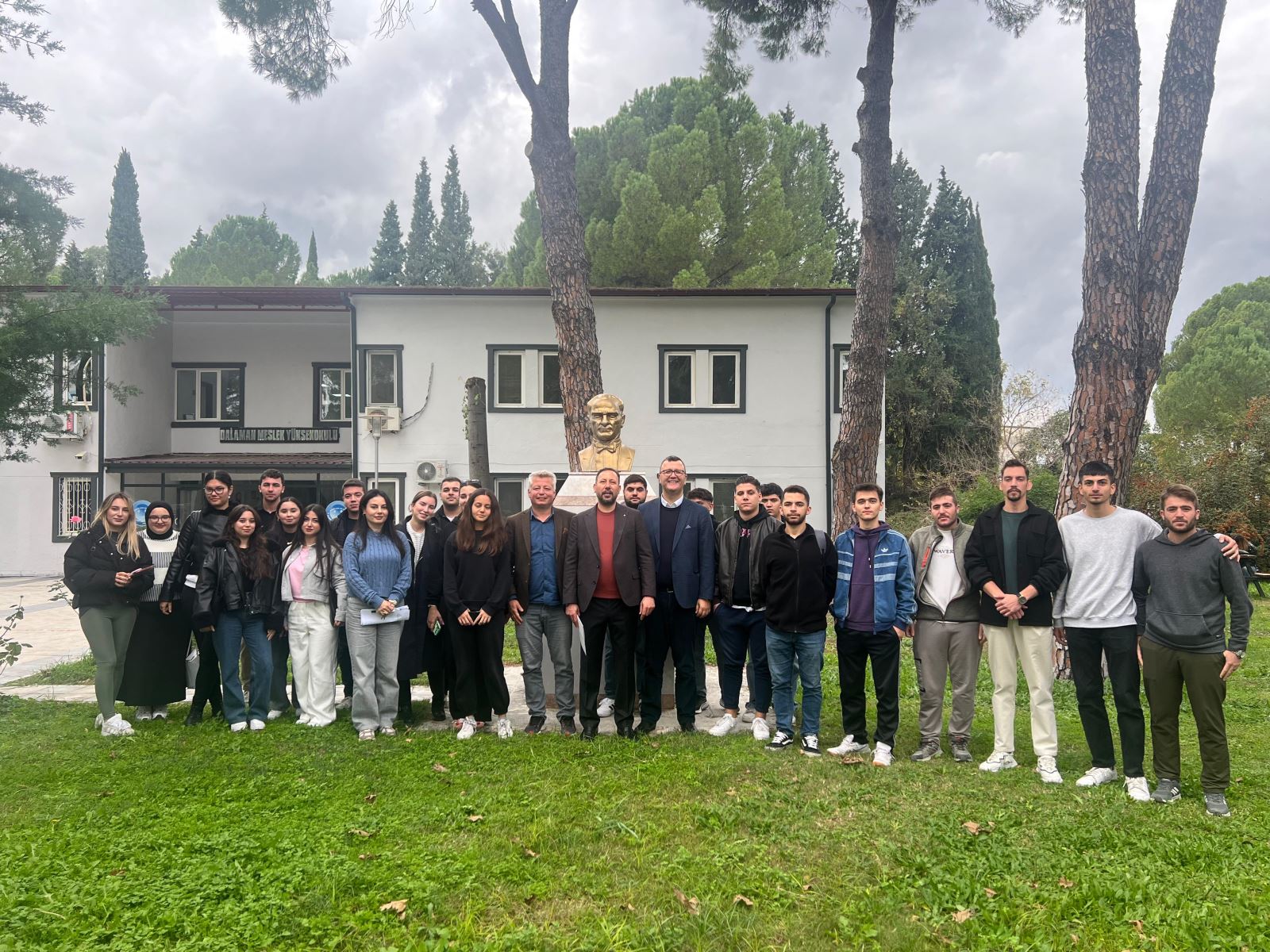 A conference held in a bright and spacious classroom at Dalaman Vocational School. Students seated at their desks are attentively listening to the speaker. A faculty member stands at the front of the room addressing the audience. Natural light enters through the windows; the classroom is full and orderly.